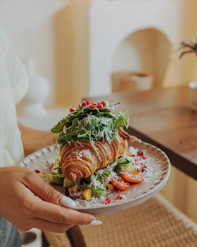 Hand hält einen Teller mit belegtem Croissant, Rucola, Avocado und Parmesan