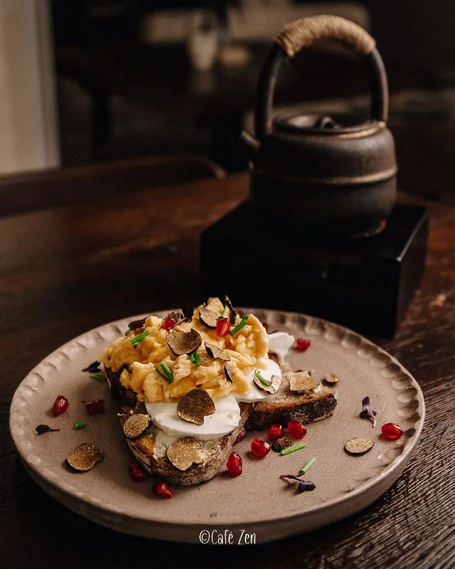 Rührei mit Trüffelscheiben auf Sauerteigbrot, dahinter eine gusseiserne Teekanne