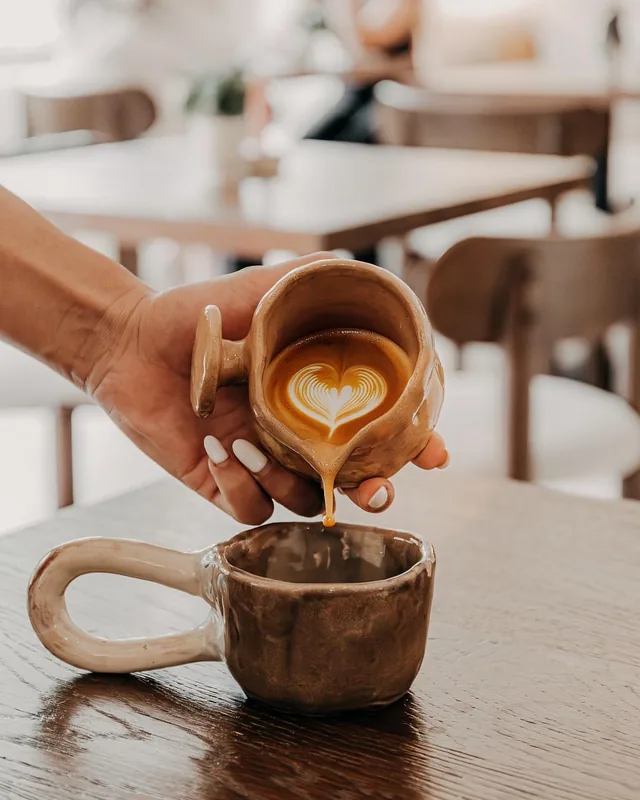 Kaffee wird aus einem Keramikkännchen in eine Tasse mit Milchschaum gegossen